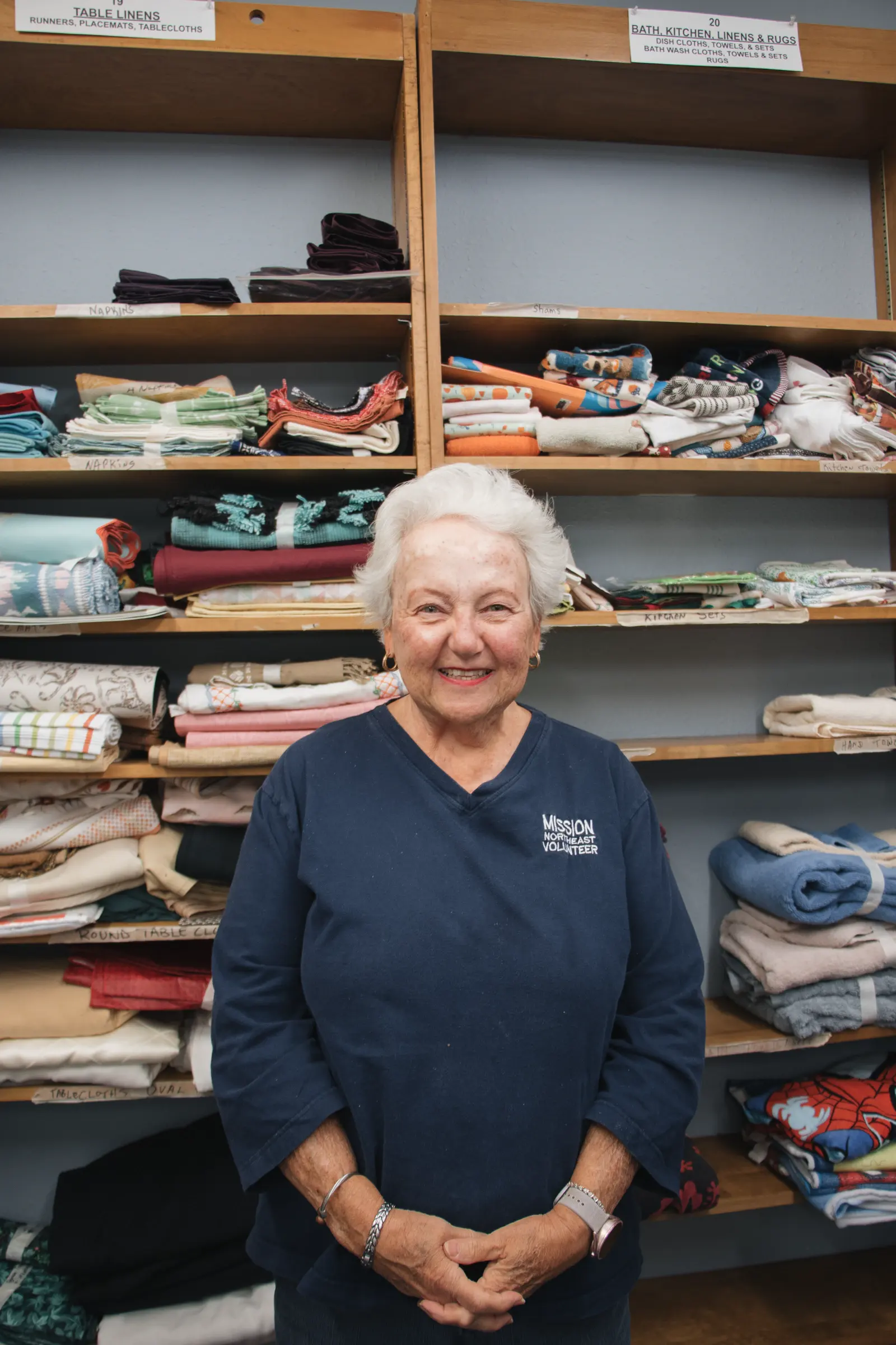 A volunteer stands in the clothing closet in front of folded linens.