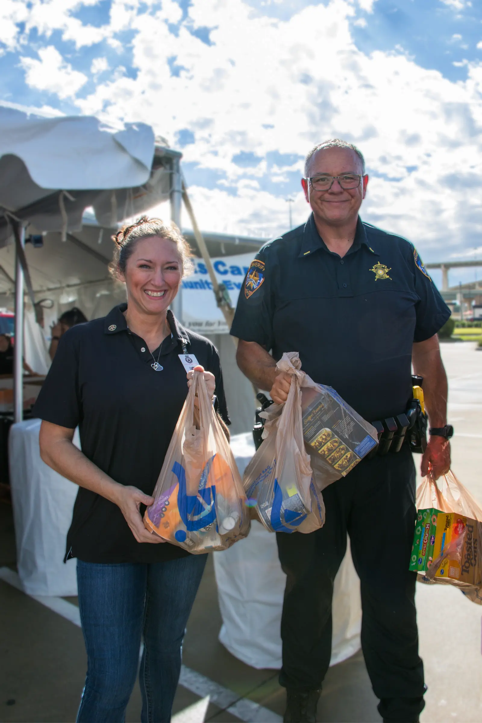 A volunteer and a sheriff’s deputy hold bags of groceries under a tent.